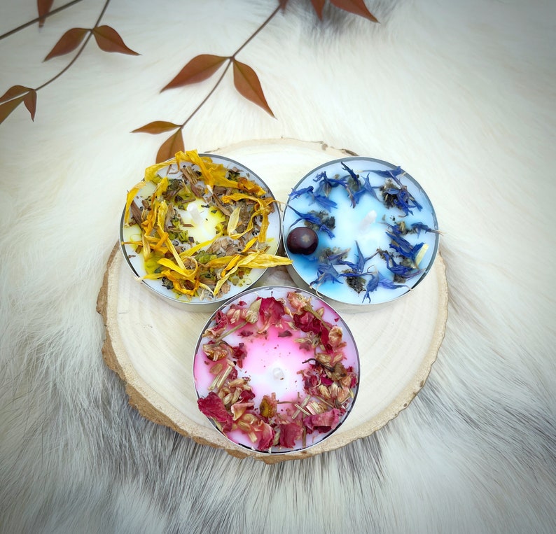 May include: Three scented tea light candles on a wooden slice. One candle has yellow petals, another has blue petals, and the third has pink petals. The candles are on a white fur-like surface.