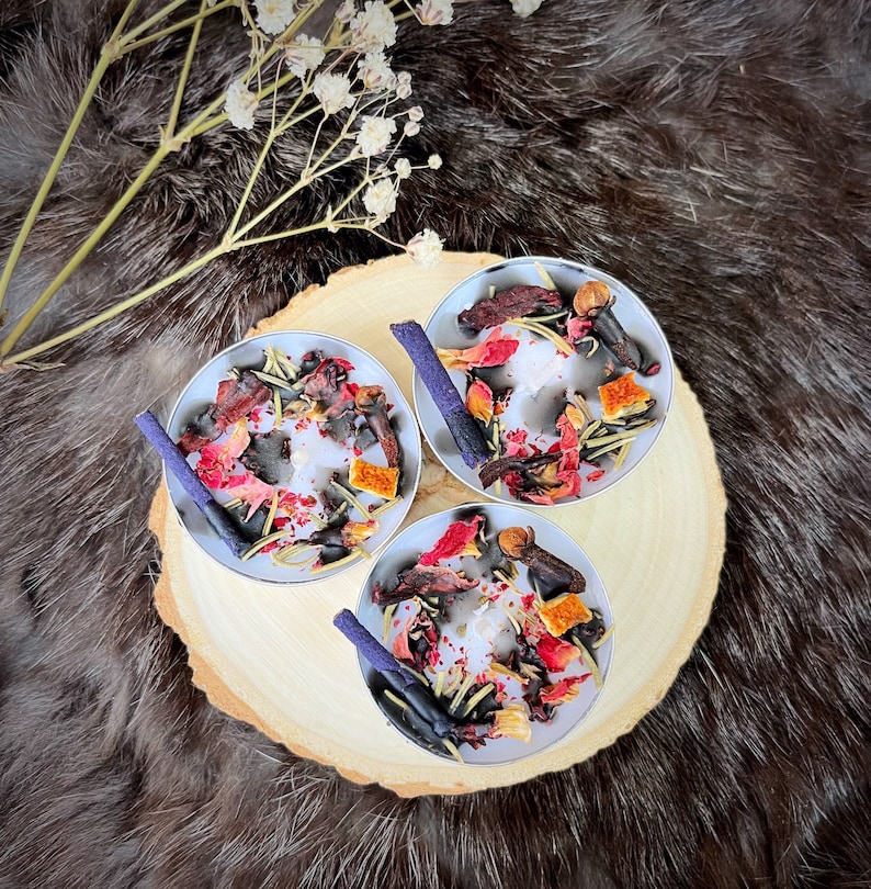 May include: Three small, round candles in silver tins, each adorned with dried flowers, spices, and a purple stick. The candles sit on a wooden slice, with a fur-like background and sprigs of white flowers.