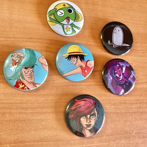 Può includere: Una collezione di sette bottoni rotondi con disegni colorati di personaggi di anime e cartoni animati. I bottoni presentano vari personaggi con colori vivaci e illustrazioni dettagliate, su una superficie di legno marrone chiaro.