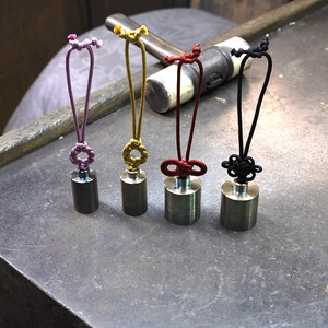 K&ouml;nnte beinhalten: F&uuml;nf kleine, zylindrische Metallgewichte mit verschiedenen farbigen Schn&uuml;ren. Die Schn&uuml;re sind in dekorativen Knoten gebunden und haben oben eine Schlaufe. Die Gewichte sind in einer Reihe auf einer grauen Oberfl&auml;che angeordnet.