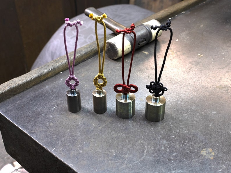 K&ouml;nnte beinhalten: F&uuml;nf kleine, zylindrische Metallgewichte mit verschiedenen farbigen Schn&uuml;ren. Die Schn&uuml;re sind in dekorativen Knoten gebunden und haben oben eine Schlaufe. Die Gewichte sind in einer Reihe auf einer grauen Oberfl&auml;che angeordnet.