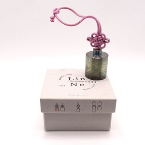 K&ouml;nnte beinhalten: Ein kleines, zylindrisches Metallgewicht mit einer strukturierten Oberfl&auml;che. Es ist an einer rosa Schnur mit einem dekorativen Knoten oben befestigt. Das Gewicht liegt auf einer wei&szlig;en Box mit dem aufgedruckten Text "Lin Ne" und "lin-ne.com".