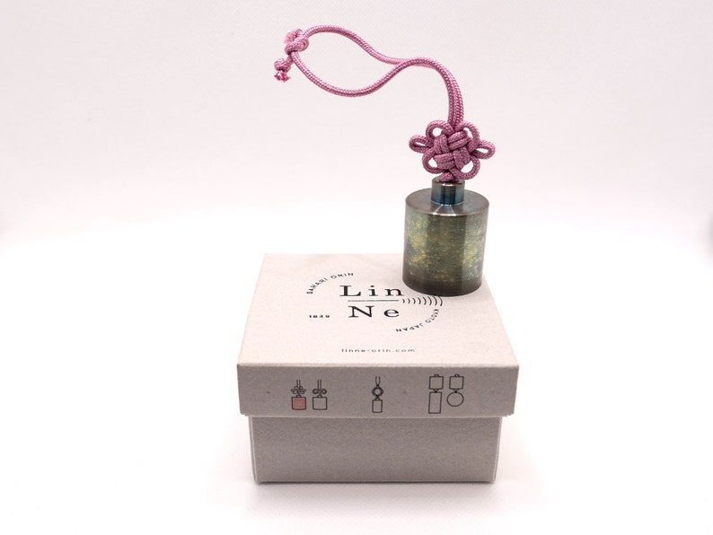 K&ouml;nnte beinhalten: Ein kleines, zylindrisches Metallgewicht mit einer strukturierten Oberfl&auml;che. Es ist an einer rosa Schnur mit einem dekorativen Knoten oben befestigt. Das Gewicht liegt auf einer wei&szlig;en Box mit dem aufgedruckten Text "Lin Ne" und "lin-ne.com".