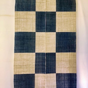 May include: A rectangular, checkered fabric panel in navy blue and beige, hanging from a wooden rod. The pattern is a grid of alternating squares. The fabric has a textured appearance, and the panel is suspended against a white background.