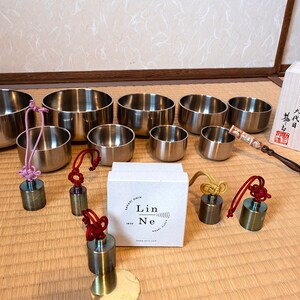 K&ouml;nnte beinhalten: Ein Satz von acht silbernen Klangschalen mit Holzschl&auml;geln und einer Schachtel mit japanischem Text. Die Schalen sind in einem Halbkreis auf einer Strohmatte angeordnet. Jede Schale hat ein kleines, rundes Messinggewicht, das an einer Schnur mit einem Knoten am Ende befestigt ist.