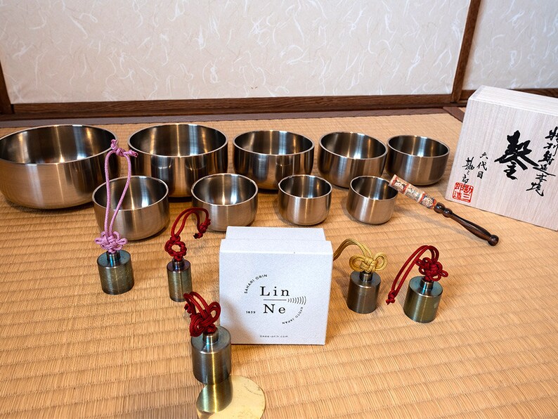 K&ouml;nnte beinhalten: Ein Satz von acht silbernen Klangschalen mit Holzschl&auml;geln und einer Schachtel mit japanischem Text. Die Schalen sind in einem Halbkreis auf einer Strohmatte angeordnet. Jede Schale hat ein kleines, rundes Messinggewicht, das an einer Schnur mit einem Knoten am Ende befestigt ist.