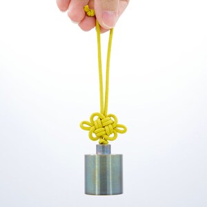 K&ouml;nnte beinhalten: Ein silbernes Metallgewicht mit einer daran befestigten gelben, geknoteten Schnur. Die Schnur ist zu einem dekorativen Knoten gebunden.