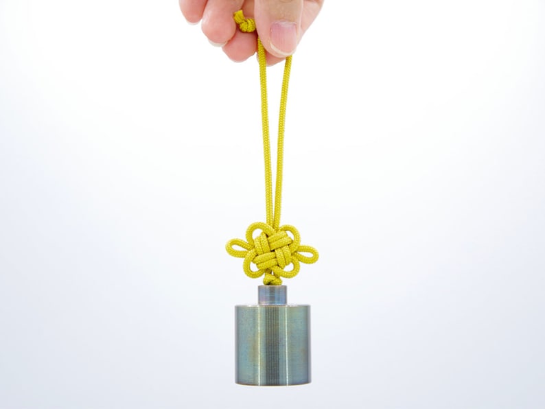 K&ouml;nnte beinhalten: Ein silbernes Metallgewicht mit einer daran befestigten gelben, geknoteten Schnur. Die Schnur ist zu einem dekorativen Knoten gebunden.