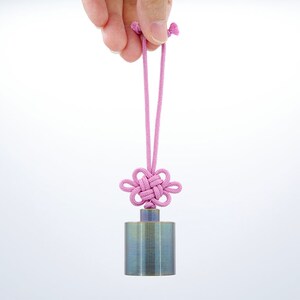 K&ouml;nnte beinhalten: Ein kleines, zylindrisches Metallgewicht mit geb&uuml;rsteter Oberfl&auml;che h&auml;ngt an einer rosa Schnur mit einem gekn&uuml;pften Blumendesign.