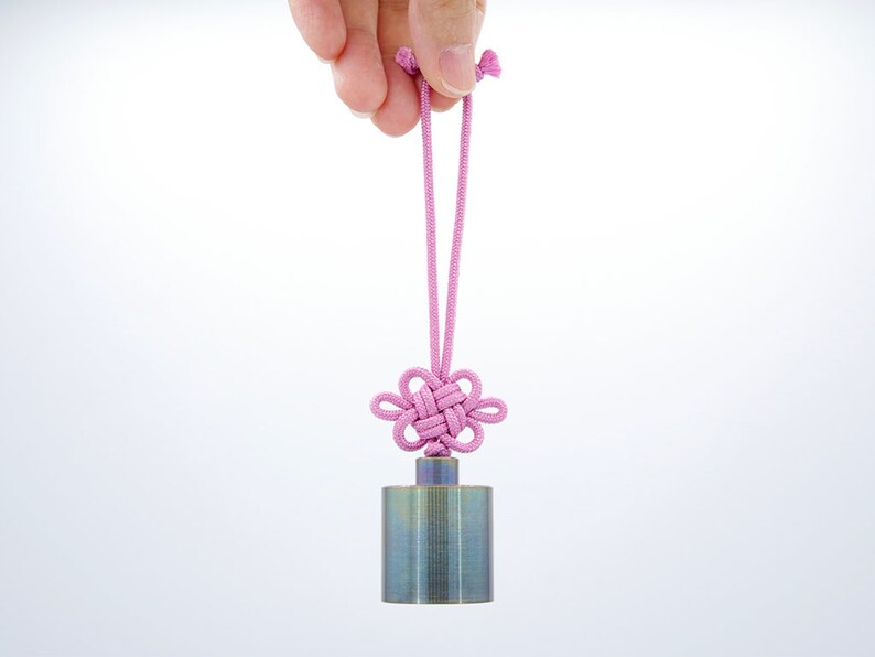 K&ouml;nnte beinhalten: Ein kleines, zylindrisches Metallgewicht mit geb&uuml;rsteter Oberfl&auml;che h&auml;ngt an einer rosa Schnur mit einem gekn&uuml;pften Blumendesign.