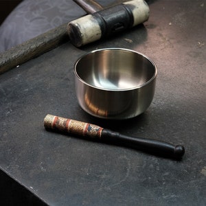 以下が含まれることがあります： 暗い表面に、銀色の歌うボウル、黒い木製のストライカー、そしてマレットがあります。ストライカーには、赤、金、茶色の模様が入った装飾的なバンドが付いています。ボウルは丸く、反射性があります。