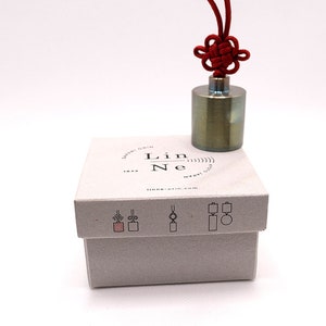 K&ouml;nnte beinhalten: Ein kleines, zylindrisches Gewicht aus gr&uuml;nem Metall mit einer roten Schnur, die in einem Knoten darum gebunden ist. Das Gewicht befindet sich in einer wei&szlig;en Box mit dem Text "Lin Ne" und einer aufgedruckten Website-Adresse.