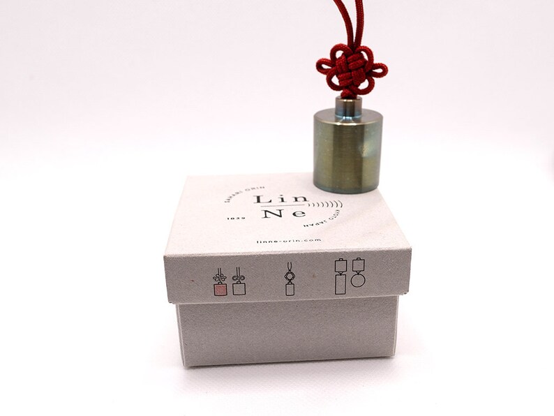 K&ouml;nnte beinhalten: Ein kleines, zylindrisches Gewicht aus gr&uuml;nem Metall mit einer roten Schnur, die in einem Knoten darum gebunden ist. Das Gewicht befindet sich in einer wei&szlig;en Box mit dem Text "Lin Ne" und einer aufgedruckten Website-Adresse.