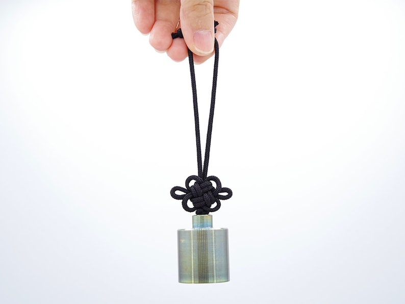 K&ouml;nnte beinhalten: Ein silberner Metallzylinder mit einem schwarzen Band, das zu einem Knoten gebunden ist, h&auml;ngt an einer Hand. Der Zylinder hat eine strukturierte Oberfl&auml;che.