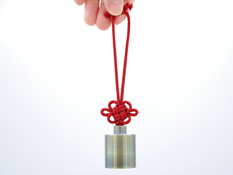 K&ouml;nnte beinhalten: Ein silbernes zylindrisches Gewicht mit einer daran befestigten roten Schnur. Der Knoten ist ein traditioneller chinesischer Gl&uuml;cksknoten.