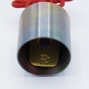 K&ouml;nnte beinhalten: Nahaufnahme eines silbernen Metallzylinders mit einer roten Schnur. Der Zylinder hat eine goldfarbene Platte im Inneren mit japanischen Schriftzeichen.