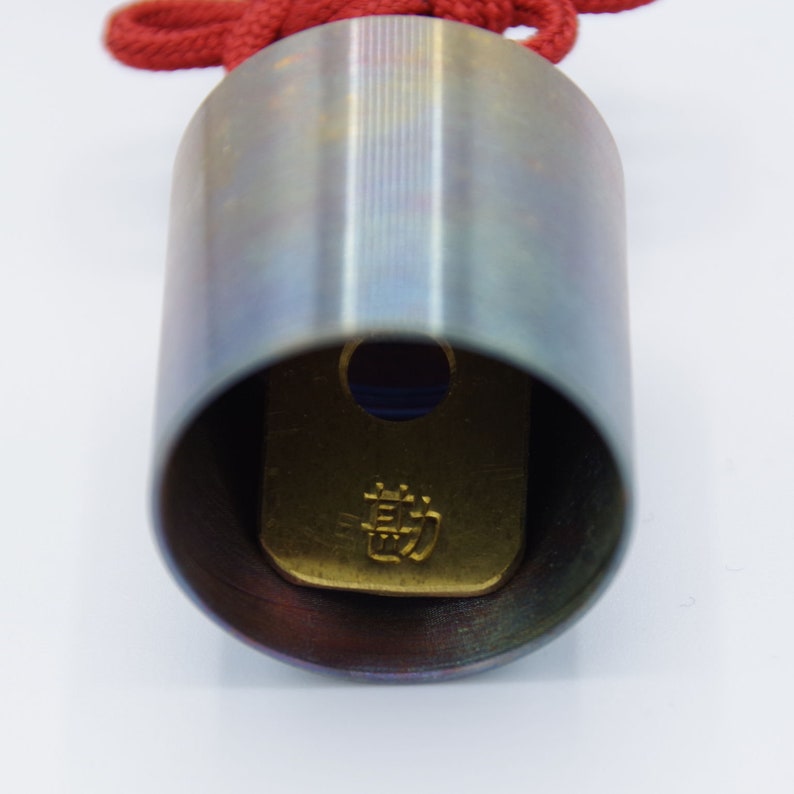 K&ouml;nnte beinhalten: Nahaufnahme eines silbernen Metallzylinders mit einer roten Schnur. Der Zylinder hat eine goldfarbene Platte im Inneren mit japanischen Schriftzeichen.