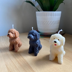 May include: Three dog-shaped candles, one brown, one black, and one white, sitting on a wooden surface.