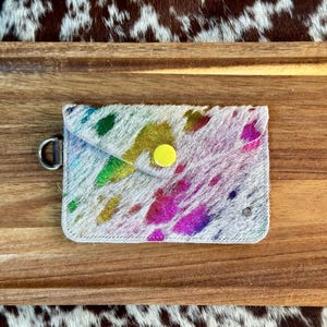 May include: A small, white leather wallet with a rainbow splatter paint design. The wallet has a yellow snap closure and a silver ring.