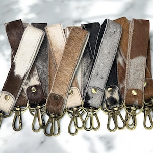 May include: A collection of leather keychains in various colors and patterns. The keychains feature a cowhide texture with brass-colored clasps and rivets. The color palette includes shades of brown, black, white, and gray.