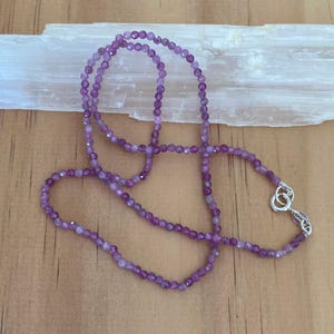 May include: A purple gemstone necklace with a silver clasp. The necklace is made of small, round beads and is approximately 18 inches long.