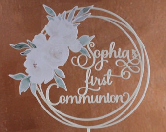 May include: White cake topper with a floral design and the text "Sophia's First Communion" in a circle.