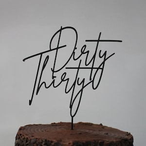 Op de afbeelding: Een chocoladetaart met een zwarte metalen taarttopper met de tekst "Dirty Thirty" in een cursief lettertype. De taart staat op een houten serveerplank. De taart heeft chocoladeschaafsel aan de zijkanten.