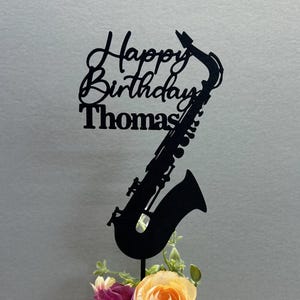 Pode incluir: Silhueta preta de um saxofone com o texto "Happy Birthday Thomas" em um fundo branco. O saxofone está colocado em um vaso branco com flores rosa e laranja.