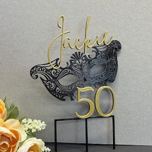 May include: Black and gold cake topper with a masquerade mask and the number 50. The topper also includes the name "Jackie" in gold script.
