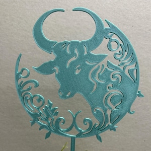 May include: A teal metal Taurus zodiac sign decoration. The bull is set within a crescent moon shape with decorative flourishes. The decoration is mounted on a teal stake and placed in a white textured pot filled with moss and small white flowers.