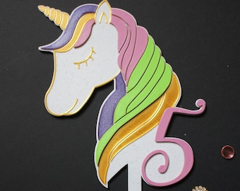 Op de afbeelding: Een witte unicorn taarttopper met een roze, groene en paarse manen en een gouden hoorn. De unicorn houdt een roze nummer "5" in zijn hoef.