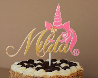 Op de afbeelding: Een witte en chocoladetaart met een roze en gouden eenhoorn topper waarop "Milda" staat.