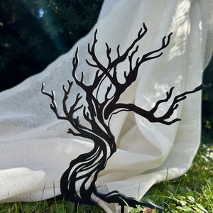 Könnte beinhalten: Eine schwarze Metallsilhouette eines Baumes mit gewundenen Ästen und einem dicken Stamm. Der Baum steht auf einem Bett aus grünem Gras mit einem weißen Tuch, das dahinter drapiert ist.