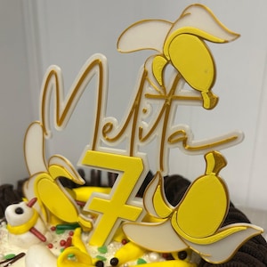 Peut inclure: Un gâteau au chocolat décoré de glaçage blanc, de décorations jaunes en forme de banane et d'un dessus blanc et or qui dit "Melita 7".