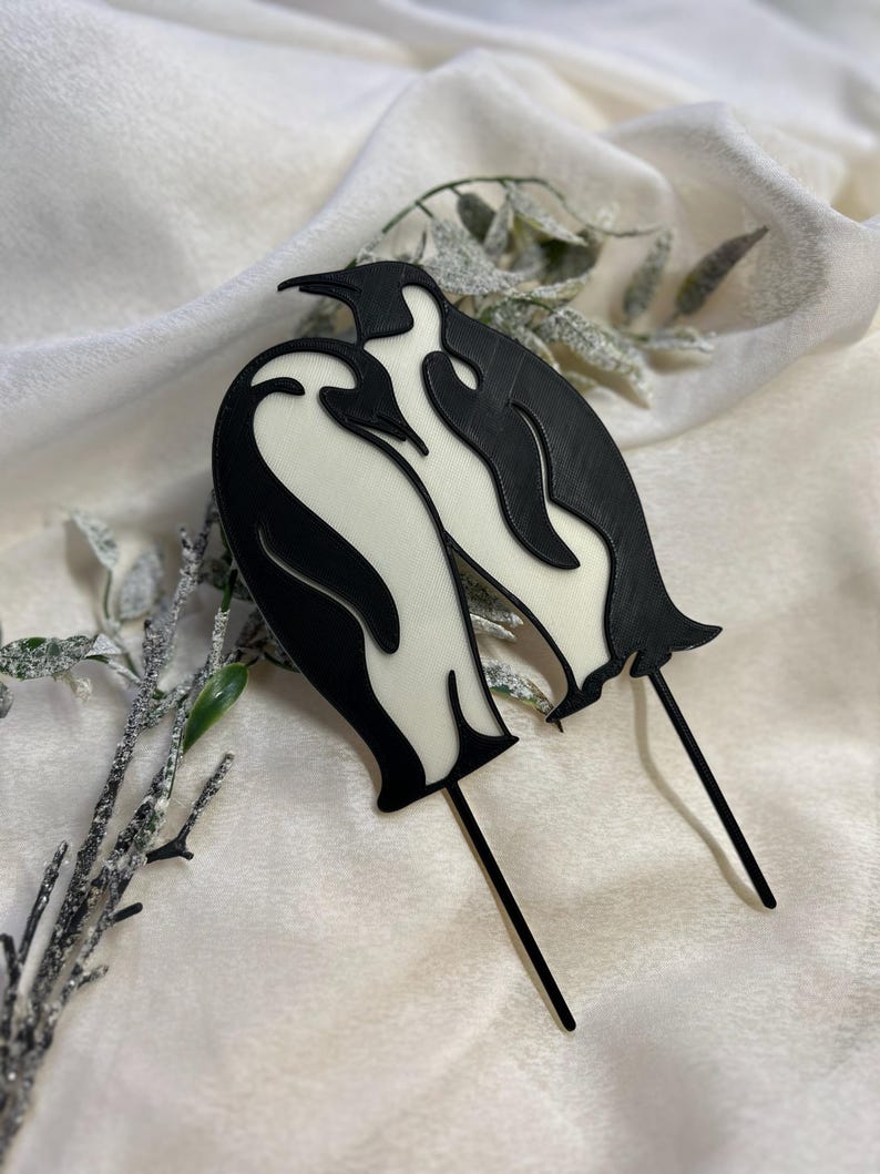 May include: Black and white penguin cake topper featuring two penguins standing together. The penguins are made of plastic and have a glossy finish.