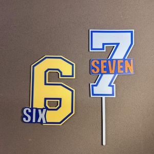 Puede incluir: Dos adornos para tartas de cumpleaños. Uno es un número 6 amarillo con un contorno azul y la palabra "SIX" en blanco. El otro es un número 7 blanco con un contorno azul y la palabra "SEVEN" en naranja, sujeto a un palito blanco.