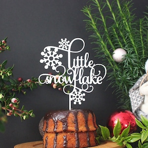 May include: White cake topper with the words "little snowflake" in cursive script. The topper is decorated with snowflakes and is sitting on top of a chocolate cake.