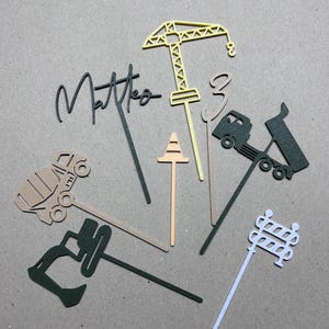 Op de afbeelding: Een verzameling cake toppers met bouwthema in verschillende kleuren. Inclusief een kraan, betonmolen, dumper en een bordje met de tekst "Matteo". De toppers zijn gemaakt van papier en zijn ontworpen voor een verjaardagstaart.