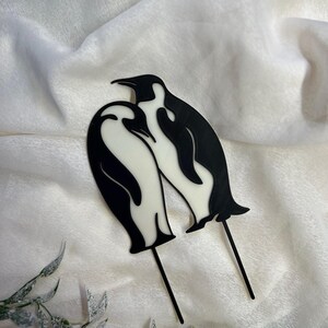 May include: Two black and white penguin silhouettes on sticks, facing each other. The penguins are in a romantic pose, with their heads tilted towards each other.