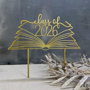 以下が含まれることがあります： 「class of 2026」の文字が入った、開いた本の形をした金色のケーキトッパー。ケーキに挿入するための細い棒が2本付いています。背景には装飾的な銀色の葉が見えます。卒業のお祝いにぴったりのエレガントなデザインです。