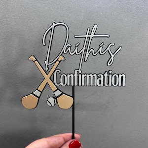 Può includere: Un cake topper con le parole "Darths Confirmation" in bianco e nero. Il topper presenta bastoni da hurling incrociati e una pallina in oro e bianco. Il topper è tenuto da un bastoncino nero.