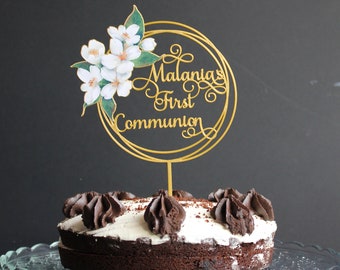 May include: A gold cake topper with white flowers and the text "Alania's First Communion".
