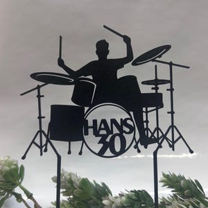 Op de afbeelding: Zwart silhouet taarttopper met een drummer die een drumstel bespeelt. De tekst "HANS 30" staat in een cirkel op de trommel. De topper is ontworpen om in een taart te worden gestoken. De achtergrond is lichtgrijs.