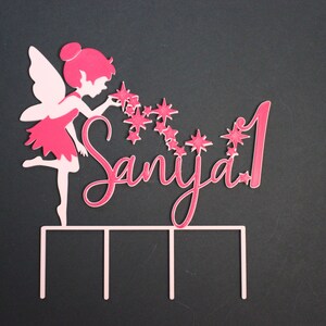 Op de afbeelding: Roze fee taarttopper met een meisje dat sterren blaast en de naam "Saniya" met het nummer "1" op een zwarte achtergrond.