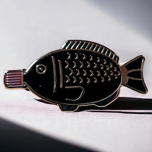 Könnte beinhalten: Eine schwarze und goldene Emaille-Anstecknadel in Form eines Fisches mit einem rosa Mund. Der Fisch hat ein detailliertes Design mit Schuppen und einer Flosse.