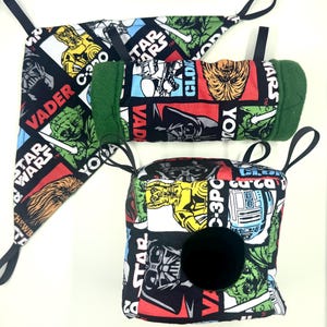 May include: Three Star Wars themed fabric items. The items include a triangular bib, a cylindrical item with green trim, and a square item with a black circle. The fabric features characters like Darth Vader, Yoda, and C-3PO.