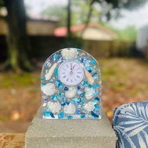 May include: A decorative clock with a clear resin arch, embedded with seashells and blue and white glass pieces. The clock face is white with black Roman numerals and black hands. The clock is sitting on a gray concrete block.