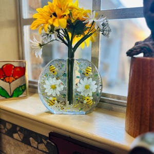 May include: A clear resin vase with a circular design, featuring yellow and black bee accents and white daisy flowers. The vase holds a bouquet of yellow sunflowers and white daisies, placed on a windowsill.