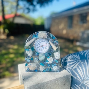 May include: A clear resin clock with an arched top, embedded with seashells and blue and white stones. The clock face is white with black Roman numerals and red hands. The clock is displayed on a gray concrete block.