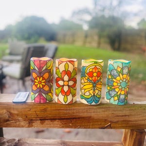 May include: Four decorative LED candles with stained-glass effect floral patterns. They are displayed on a wooden surface, with a remote control. The background is a blurred outdoor scene with greenery and seating.